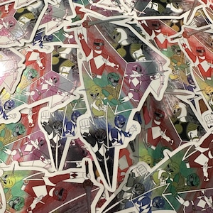 Pode incluir: Uma pilha de adesivos coloridos com personagens dos Power Rangers em várias poses de ação. Os adesivos mostram os Rangers em suas cores características: vermelho, azul, verde, amarelo, rosa e preto, com armas e capacetes.