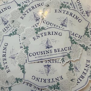 Op de afbeelding: Witte stickers met een blauw en groen ontwerp. De stickers tonen de tekst "Entering Cousins Beach" met een zeilboot illustratie en bloemaccenten. De zin "My whole life was measured in summers" is ook aanwezig.
