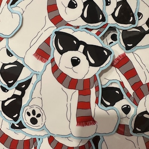 May include: A collection of white bear stickers, each wearing black sunglasses and a red and gray striped scarf. The stickers have a light blue outline and a paw print on one paw.