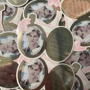 Op de afbeelding: Ovale stickers met een portret van een jonge man omringd door witte bloemen. De stickers hebben een gouden rand en een roze lint. De tekst op de sticker luidt: "for belly, conrad is the sun, and when the sun comes out, the stars disappear."