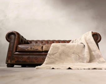 May include: A brown leather Chesterfield sofa with a tufted back and rolled arms. A cream-colored faux fur throw blanket is draped over the sofa, adding a touch of luxury to the living room decor.