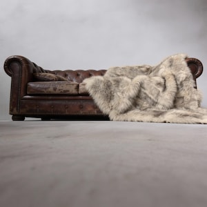 Puede incluir: Un sofá Chesterfield de cuero marrón con respaldo capitoné y brazos enrollados. Una manta de piel sintética en tonos grises y beige está sobre el sofá. El entorno es una habitación con suelo y pared gris neutro.