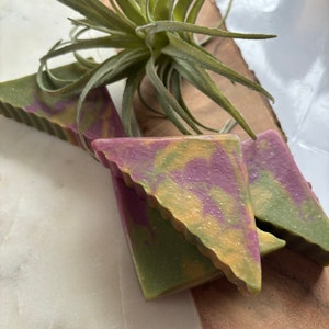 Könnte beinhalten: Drei dreieckige Seifenstücke mit einem grünen, gelben und violetten Wirbelmuster. Die Seife liegt auf einer hellbraunen Oberfläche.