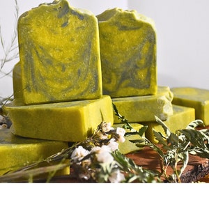 May include: A stack of handmade soap bars with a vibrant green hue and dark gray marbling. The rectangular bars are arranged on a wooden surface, with sprigs of greenery and white flowers adding a natural touch. The soap appears to be freshly made.