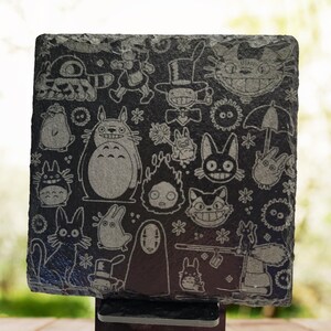 May include: A black slate coaster with a white engraved design featuring characters from the animated film "My Neighbor Totoro". The coaster is on a black stand. "Style 3" is written in white on a wooden surface.