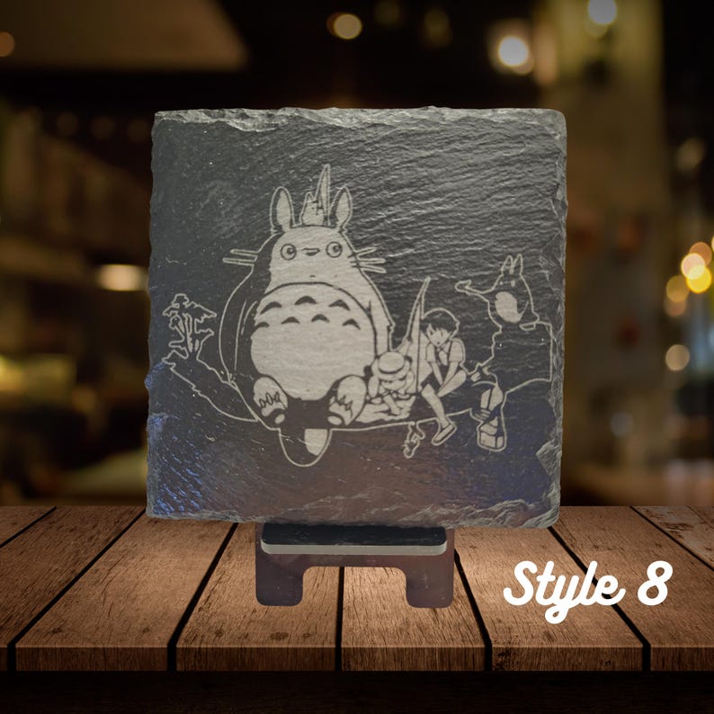 May include: A black slate coaster with a white etched image of Totoro and two characters from the movie My Neighbor Totoro. The coaster is on a wooden stand. Style 8.