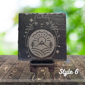 May include: A black slate coaster with a white engraved design of a cartoon character with a mountain and sun inside. The coaster is on a wooden stand. Style 6.