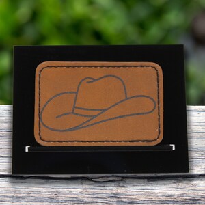 May include: A brown leather patch with a black outline of a cowboy hat.