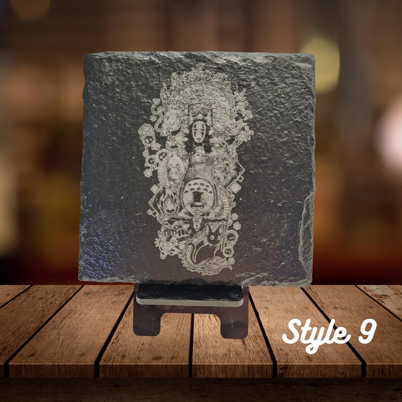 May include: A black slate coaster with a white engraved image of characters from the animated film Spirited Away. The coaster is on a wooden stand. Style 9.