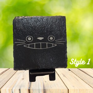 May include: Black slate coaster with a white engraved cartoon face of a cat with large eyes and a wide smile. The coaster is on a wooden stand. "Style 1" is written in white text on the right side of the image.