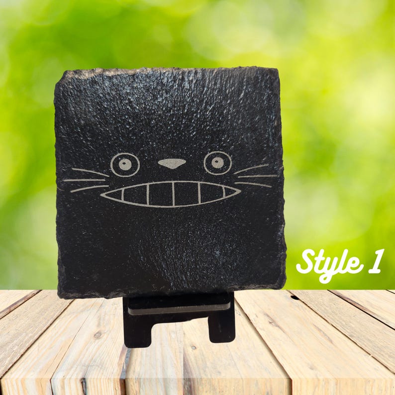 May include: Black slate coaster with a white engraved cartoon face of a cat with large eyes and a wide smile. The coaster is on a wooden stand. "Style 1" is written in white text on the right side of the image.