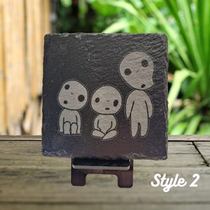 May include: A black slate coaster with a white design of three cartoon characters. The characters are from the movie "My Neighbor Totoro". The coaster is on a wooden stand. The text "Style 2" is written in white on a black background.