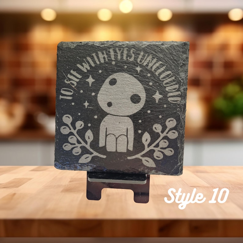 May include: A black slate coaster with a white design of a character with a mushroom head and the text "To sit with eyes unclouded". The coaster is on a wooden stand.