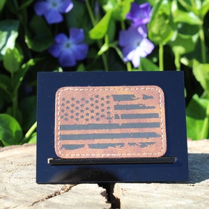 May include: A distressed leather patch with a black and white American flag design. The patch is displayed on a black stand.