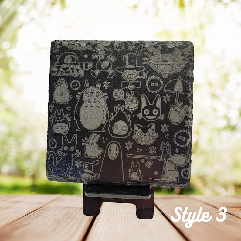 May include: A black slate coaster with a white engraved design featuring characters from the animated film "My Neighbor Totoro". The coaster is on a black stand. "Style 3" is written in white on a wooden surface.