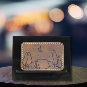 May include: A brown leather wallet with a stitched border and a black and white illustration of a tent, trees, and a crescent moon.