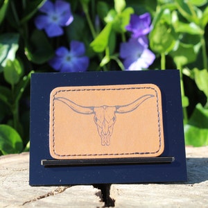Könnte beinhalten: Ein braunes Lederstück mit einem genähten Rand, das eine schwarze Linienzeichnung eines Longhorn-Schädels mit großen Hörnern zeigt. Das Stück wird auf einem schwarzen Ständer vor einem verschwommenen Hintergrund aus grünen Blättern und lila Blüten präsentiert.
