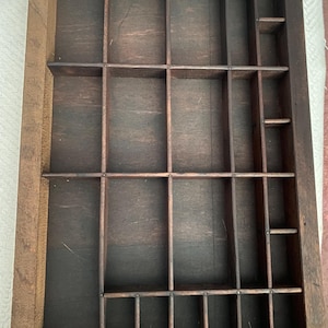 May include: A rectangular dark brown wooden printer's tray featuring multiple compartments of varying sizes separated by thin wooden dividers. The tray has a sturdy outer frame and a flat base, designed with a grid-like layout for storage.