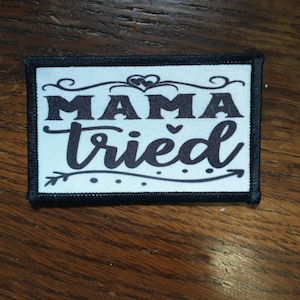 以下が含まれることがあります： 黒い刺繍で「ママは試した」と書かれた白いパッチ。ハートと矢印のデザインが施されています。