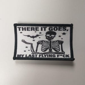 May include: Black and white embroidered patch with a skeleton sitting on a cloud with the text "THERE IT GOES, MY LAST FLYING F*CK" and stars and a bat.