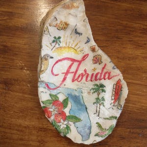 Może przedstawiać: Ozdobna muszla ostrygi z ręcznie malowaną mapą Florydy. Muszla zawiera napis "Florida" w czerwonym skrypcie, a także ilustracje zachodu słońca, palm oraz lokalnej flory i fauny.