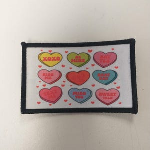 May include: A white fabric patch with a black border featuring nine colourful heart-shaped sweets with different messages, including "XOXO", "Be Mine", "Say Yes", "Kiss Me", "Text Me", "Miss You", "Best Day", and "Sweet Talk".