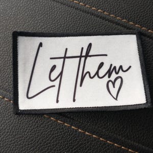 May include: White fabric patch with black stitching around the edges. The patch has black text that says "Let them" with a small black heart.