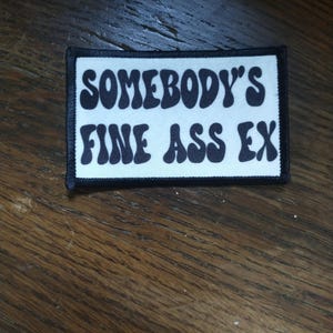 May include: A rectangular patch with a white background and a black border. The patch features the text "SOMEBODY'S FINE ASS EX" in a bold, retro font. The patch is likely designed to be ironed on or sewn onto clothing or accessories.
