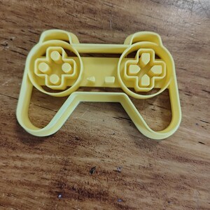 Może przedstawiać: Żółta plastikowa foremka do ciastek w kształcie kontrolera do gier wideo. Posiada dwie okrągłe sekcje z przyciskami w kształcie krzyża i centralny pasek z zakrzywioną podstawą.