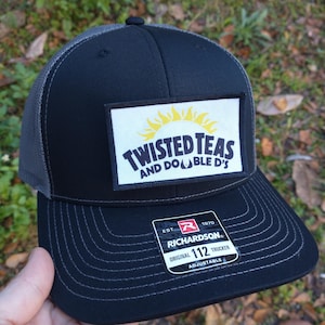 May include: Black and gray trucker hat with a yellow sun and the text "Twisted Teas and Double D's" on a white patch. The hat is adjustable and has the Richardson logo and the text "Original 112 Trucker" on a black and red patch.