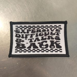 Op de afbeelding: Zwart-witte strijkpatch met de tekst "Expensive Difficult & Talks Back" in een retro-lettertype met een geruite rand.