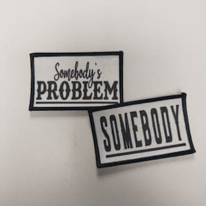 Può includere: Due toppe ricamate in bianco e nero con sfondo bianco e testo nero. La toppa superiore dice "Somebody's PROBLEM" e la toppa inferiore dice "SOMEBODY".