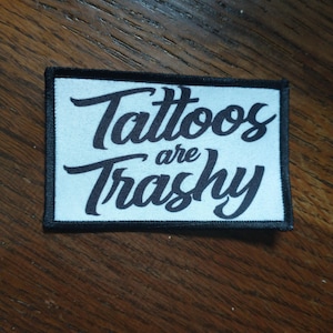 Può includere: Toppa bianca con testo nero che recita "Tattoos are Trashy" in un font stilizzato.