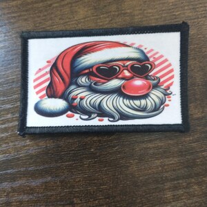 May include: A white patch with a black border features a cartoon illustration of Santa Claus wearing heart-shaped sunglasses and a red hat. The background is a red and white striped circle.
