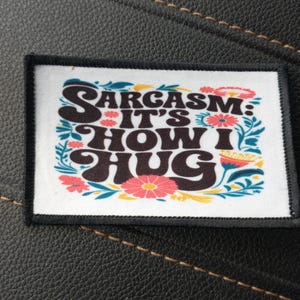 May include: A white patch with black text that reads "Sarcasm: It's How I Hug" surrounded by colourful flowers. The patch has a black border.