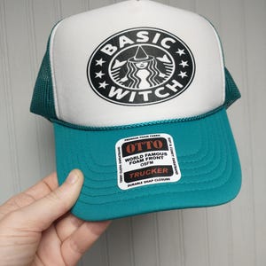 May include: A teal and white trucker hat with the words "BASIC WITCH" in a circular design. The hat has a teal brim and mesh back, with a white front panel. The hat is made of foam and has a snap closure.