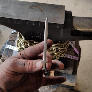 May include: A close-up of a silver metal blade being held by a hand. The blade is thin and flat, with a pointed tip. The hand is holding the blade by the handle, which is made of the same metal as the blade. The blade is resting on a wooden surface.