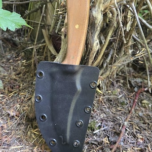 May include: A fixed-blade knife with a wooden handle and a black sheath. The handle is light brown with two brass rivets. The black sheath has multiple rivets around the edges. The knife is partially inserted into the sheath.