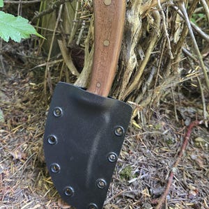 May include: A knife with a wooden handle and a black sheath. The handle is a light brown colour and is secured with two rivets. The sheath is made of a black material and has several rivets around the edge. The knife is resting in a natural outdoor setting.