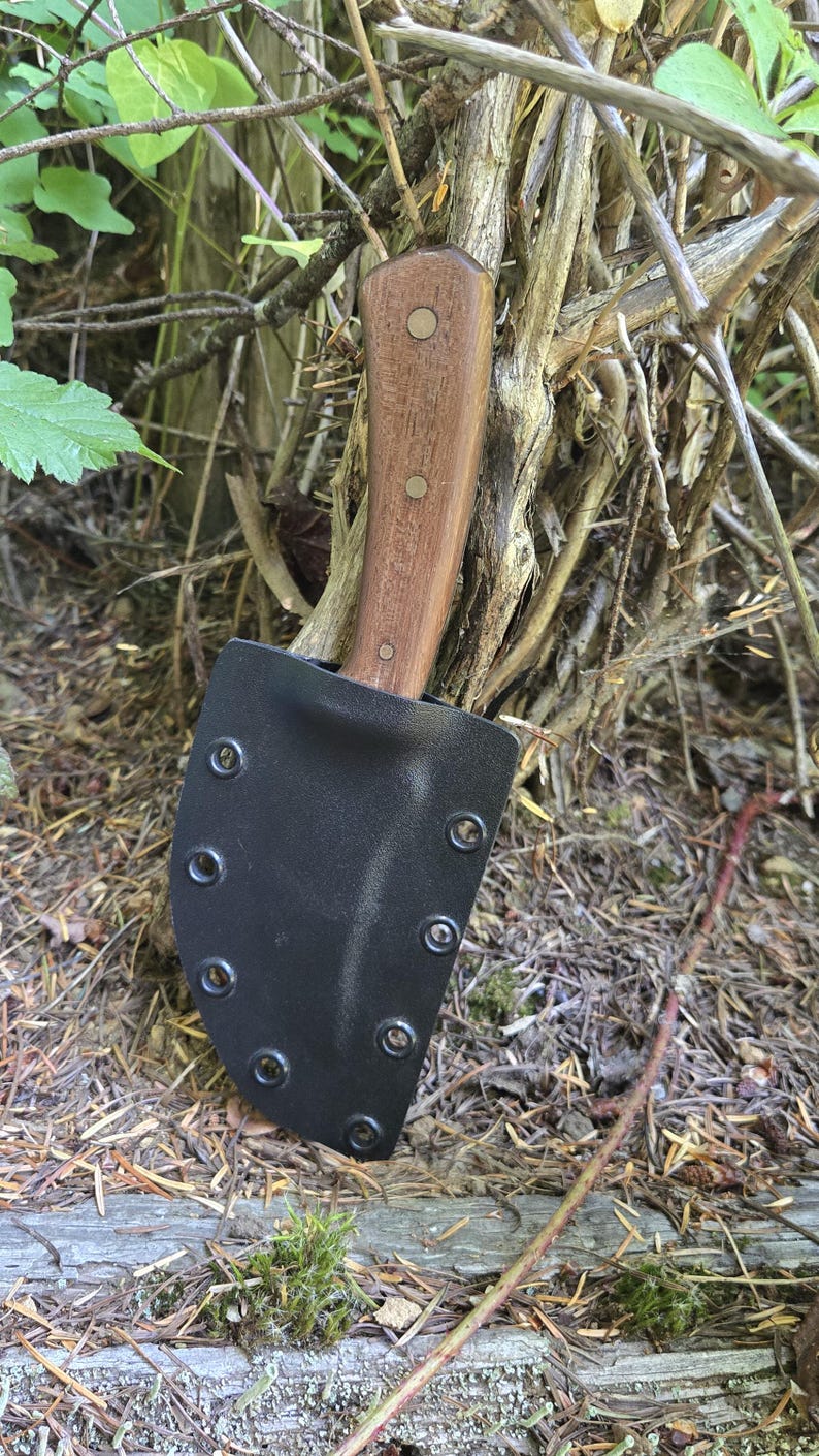 May include: A knife with a wooden handle and a black sheath. The handle is a light brown colour and is secured with two rivets. The sheath is made of a black material and has several rivets around the edge. The knife is resting in a natural outdoor setting.