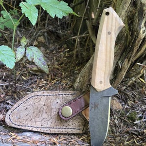 May include: A fixed-blade knife with a light wood handle and a dark blade, in a textured leather sheath with a brown leather strap and brass button. The knife is set against a natural outdoor backdrop.
