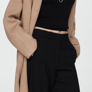 Könnte beinhalten: Eine Frau trägt einen langen, camel-farbenen Cardigan mit langen Ärmeln über einem schwarzen, figurbetonten Oberteil und einer schwarzen, weiten Hose. Sie trägt eine silberne Halskette mit einem geometrischen Anhänger.