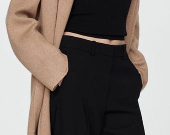 May include: A woman wearing a camel-coloured long-sleeved cardigan over a black fitted top and black wide-leg trousers. She is wearing a silver necklace with a geometric pendant.