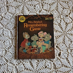 May include: A vintage copy of "Mrs. Brisby's Remembering Game," a Little Golden Sniff It Book. The cover features illustrations of cartoon mice in various colors, set against a brown background with the title in large, gold lettering. The book is placed on a cream-colored, crocheted surface.