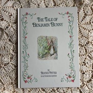 May include: A hardcover book titled "The Tale of Benjamin Bunny" by Beatrix Potter. The cover is white with green and pink floral accents. A color illustration of rabbits is in the center. An authorized edition.