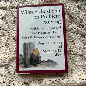 May include: A hardcover book titled "Winnie-the-Pooh on Problem Solving." The cover features an illustration of Winnie the Pooh in a suit, sitting near a tower and a laptop. The book's title and author names are in white text on a burgundy background.