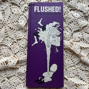 May include: A purple book titled "Flushed! The W.C. Companion" with a humorous illustration of a person being flushed down a toilet. The cover features a cartoon-style drawing in black and white, with the person's legs and arms visible.