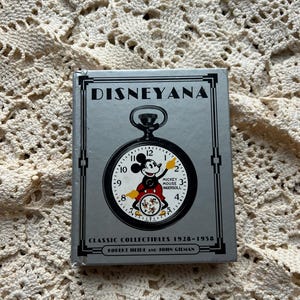 May include: A silver box titled "DISNEYANA" features a pocket watch design with Mickey Mouse. The watch face shows the time and the words "MICKEY MOUSE INGERSOLL." The box also includes the text "CLASSIC COLLECTIBLES 1928-1958" and the names Robert Heide and John Gilman.