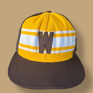 Puede incluir: Una gorra de béisbol marrón y amarilla con una visera marrón. La gorra presenta una corona amarilla con rayas blancas y una letra "W" marrón. La parte trasera de la gorra es de malla marrón.