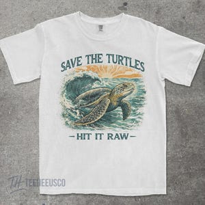 May include: White t-shirt with a graphic of a sea turtle swimming in the ocean. The text "SAVE THE TURTLES" is above the turtle, and "- HIT IT RAW -" is below. The design features shades of blue, green, and orange.